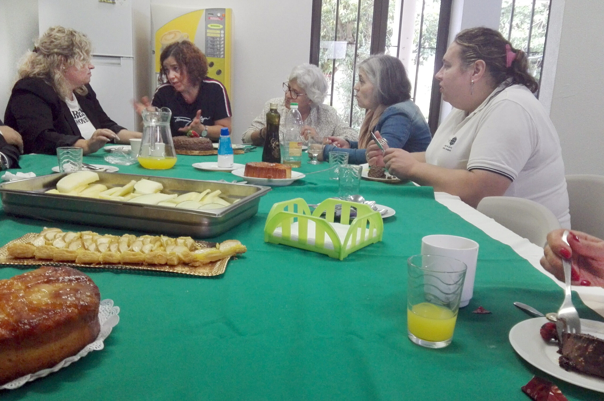 Cinco pessoas sentadas à mesa e a conversar. Em cima da mesa estão vários bolos, frutas laminadas, sumos, pratos e copos.