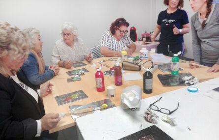 Seis pessoas junto de uma mesa na qual estão pousados materiais para elaboração de um trabalho de expressão artística.