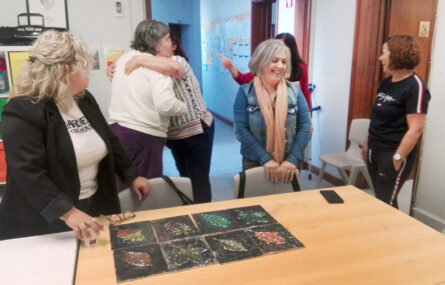 Seis pessoas junto de uma mesa na qual estão pousados oito trabalhos de expressão artística e plástica. Duas das pessoas abraçam-se.