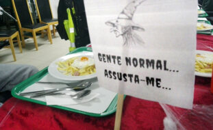 Mesa com um tabuleiro (no qual estão um prato com comida e alguns talheres). Ao fundo algumas cadeiras, vazias. Em primeiro plano um papel com a imagem de uma bruxa e a frase Gente normal assusta-me...