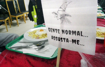 Mesa com um tabuleiro (no qual estão um prato com comida e alguns talheres). Ao fundo algumas cadeiras, vazias. Em primeiro plano um papel com a imagem de uma bruxa e a frase Gente normal assusta-me...