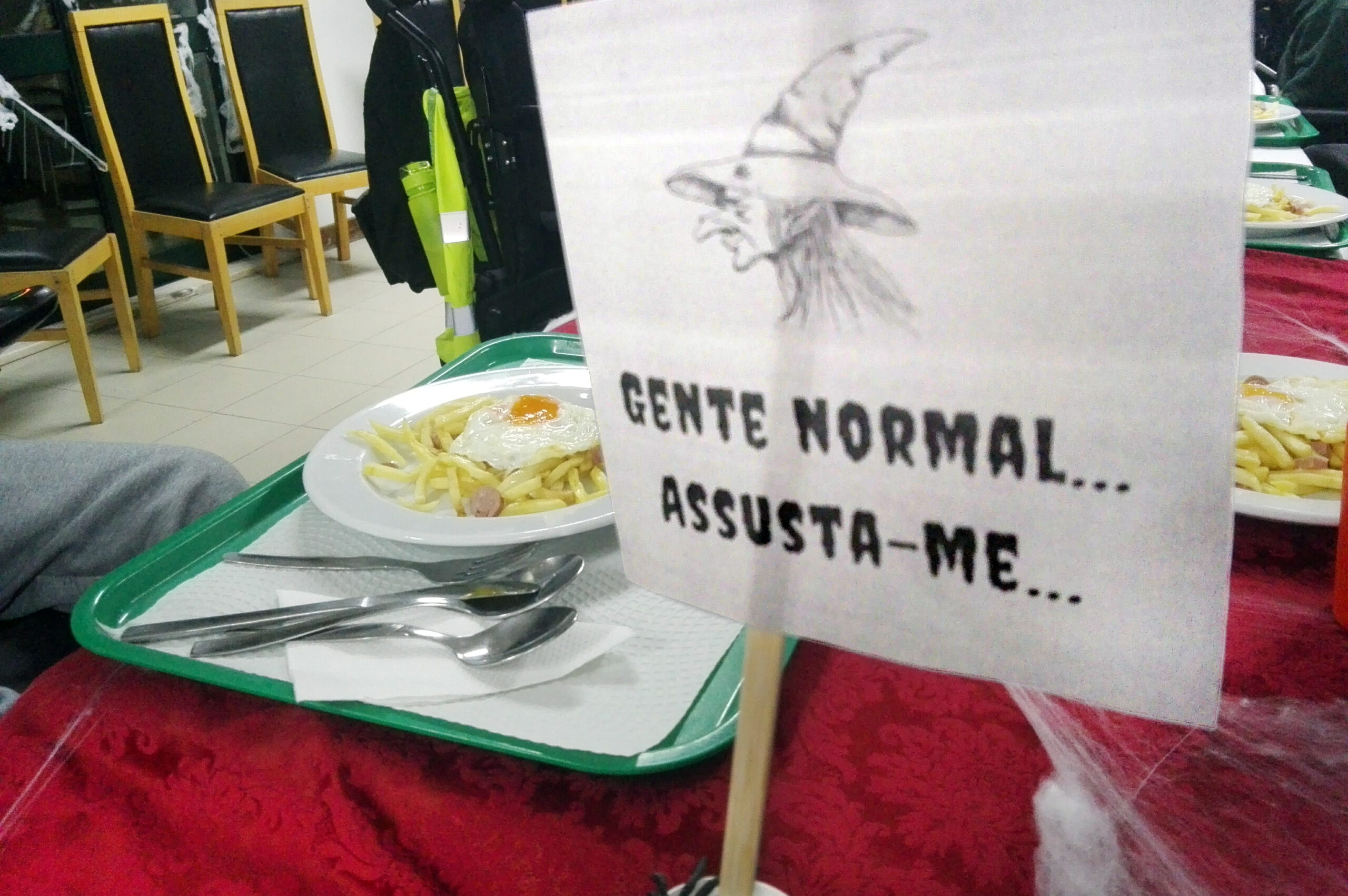 Mesa com um tabuleiro (no qual estão um prato com comida e alguns talheres). Ao fundo algumas cadeiras, vazias. Em primeiro plano um papel com a imagem de uma bruxa e a frase Gente normal assusta-me...