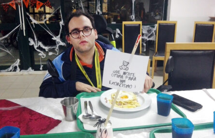 Fotografia de uma pessoa sentada à mesa. Junto do prato (com comida) e do copo (com um qualquer líquido...) está um pequeno cartaz com uma imagem de um urso e a expressão “Que noite ótima para um exorcismo”.