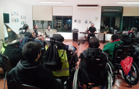 Fotografia de sala interior com uma pessoa que utiliza um órgão ou sintetizador enquanto outras duas cantam. Em primeiro plano várias pessoas a assistir e sentadas em semi-círculo.