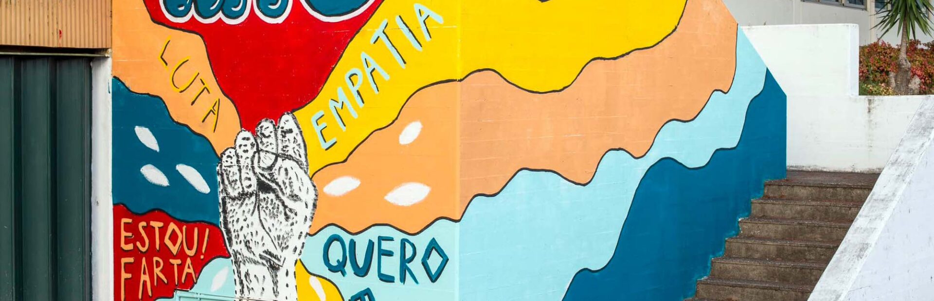 Parede do Centro de Reabilitação pintada. O desenho é constituído por um punho cerrado, a partir do qual explodem várias cores: azul, laranja, amarelo e vermelho. Em cima está escrito “APPC”. Nas cores, várias palavras surgem: “quero entrar”, “estou farta”, “empatia” e “luta”.