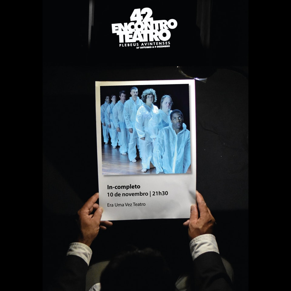 Fotografia, em plano superior de duas mãos a segurarem um cartaz. Acima do cartaz surge a indicação do 42.º Encontro de Teatro de Os Plebeus Avintenses. O cartaz é composto de uma fotografia de sete atores vestidos com roupas integralmente brancas e alinhados, em fila. Depois, logo abaixo, surge o nome da peça (IN-completo), data e hora de apresentação (10 de novembro às 21h30) e o nome da companhia (Era uma vez Teatro).