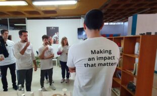 Fotografia de espaço interior com grupo de pessoas a conversar. Todas usam t-shirt branca sendo que em primeiro plano, de costas, vê-se numa delas impressa a expressão “Making an impact that matters”.