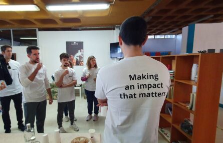 Fotografia de espaço interior com grupo de pessoas a conversar. Todas usam t-shirt branca sendo que em primeiro plano, de costas, vê-se numa delas impressa a expressão “Making an impact that matters”.