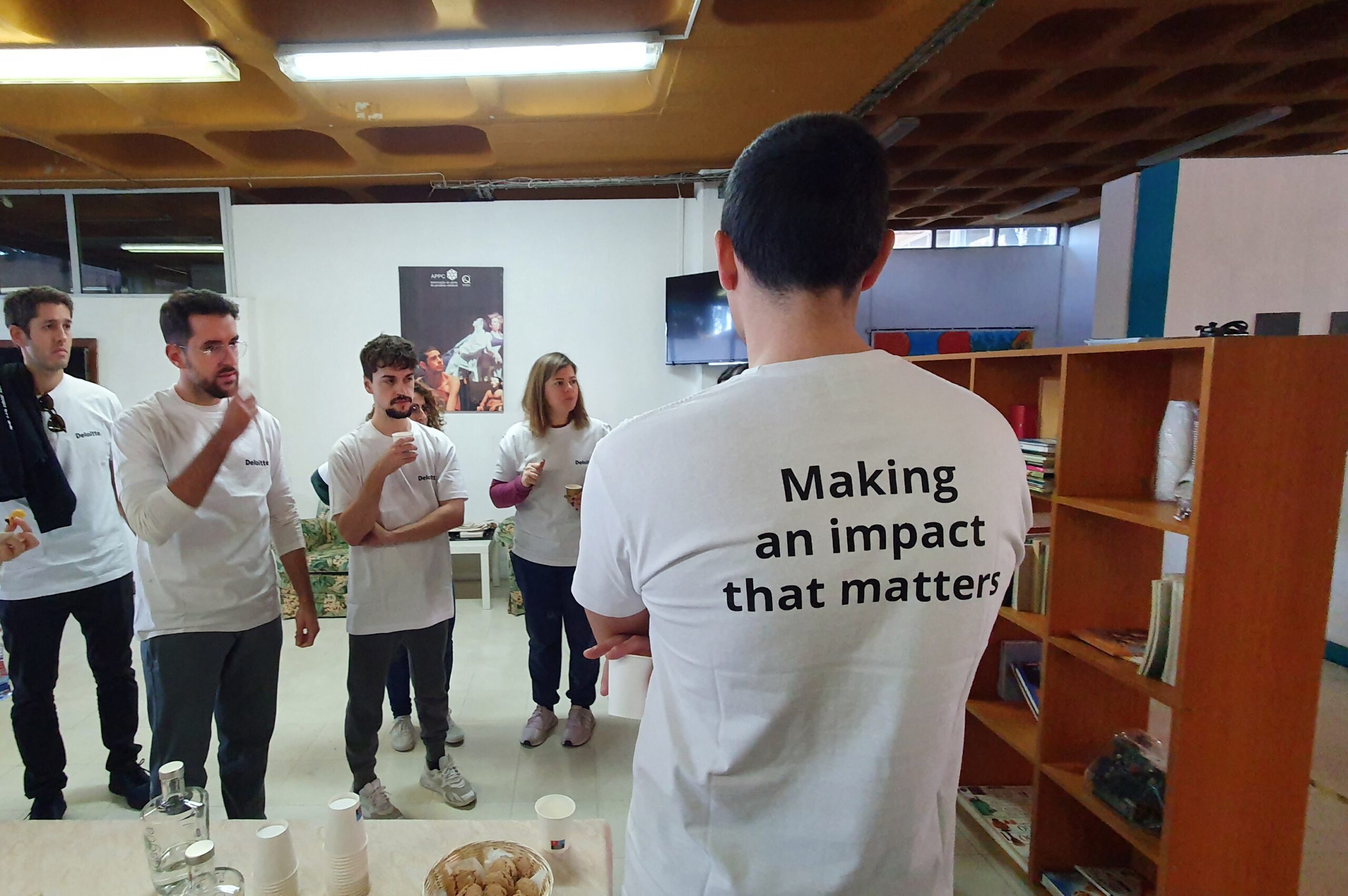 Fotografia de espaço interior com grupo de pessoas a conversar. Todas usam t-shirt branca sendo que em primeiro plano, de costas, vê-se numa delas impressa a expressão “Making an impact that matters”.