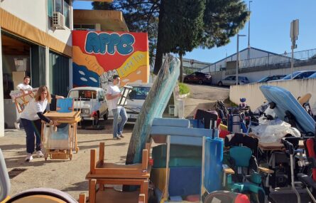 Fotografia de zona exterior de estacionamento da APPC. Em primeiro plano incontáveis produtos de apoio a serem organizados. Mais ao fundo duas viaturas estacionadas e um mural, colorido, com a APPC em destaque (a azul). Árvores ao fundo.