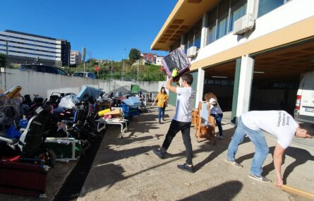 Fotografia de zona exterior de estacionamento da APPC. Do lado esquerdo estão incontáveis produtos de apoio a serem organizados. Mais à direita várias pessoas transportam e organizam alguns desses produtos de apoio.