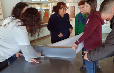 Seis pessoas junto de uma mesa que se encontra em fase de recuperação e melhoria. Estão a colocar e colar aquilo que aparenta ser uma proteção em plástico ou vinil no tampo da referida mesa.