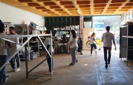 Fotografia de espaço interior (zona de garagem) da APPC. Ainda por retirar estão alguns produtos de apoio. Do lado esquerdo da imagem e ao fundo ainda são visíveis alguns desses produtos e inúmeras caixas com equipamento e material não identificável. Uma pessoa carrega material para o exterior.