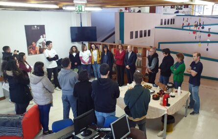 Grupo de pessoas em espaço interior da APPC, em círculo e em perspetiva superior. Do lado direito vê-se uma mesa com máquina de café, chá e alguns biscoitos. Ao fundo da imagem escadaria e rampas de acesso.