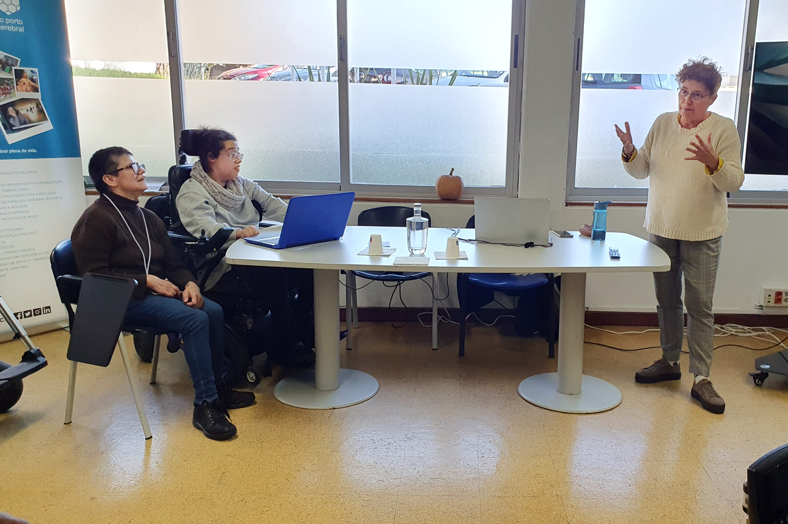 Três pessoas junto de uma mesa. À esquerda estão duas sentadas e do lado direito (da imagem) está uma pessoa em pé, aparentemente a falar e gesticular. Em cima da mesa estão dois computadores portáteis.
