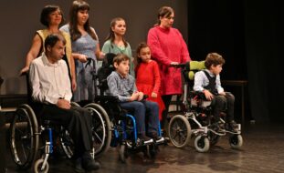 Oito pessoas em palco, sendo que três estão sentadas em cadeiras de rodas.
