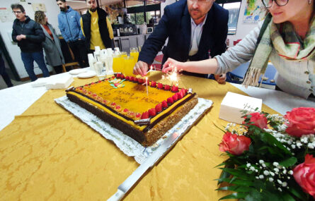 Duas pessoas acendem as velas de um bolo de aniversário. Em cima da mesa, além do bolo, está um arranjo floral, pratos, guardanapos, copos e jarros com sumo. Mais ao fundo da imagem outras quatro pessoas assistem.