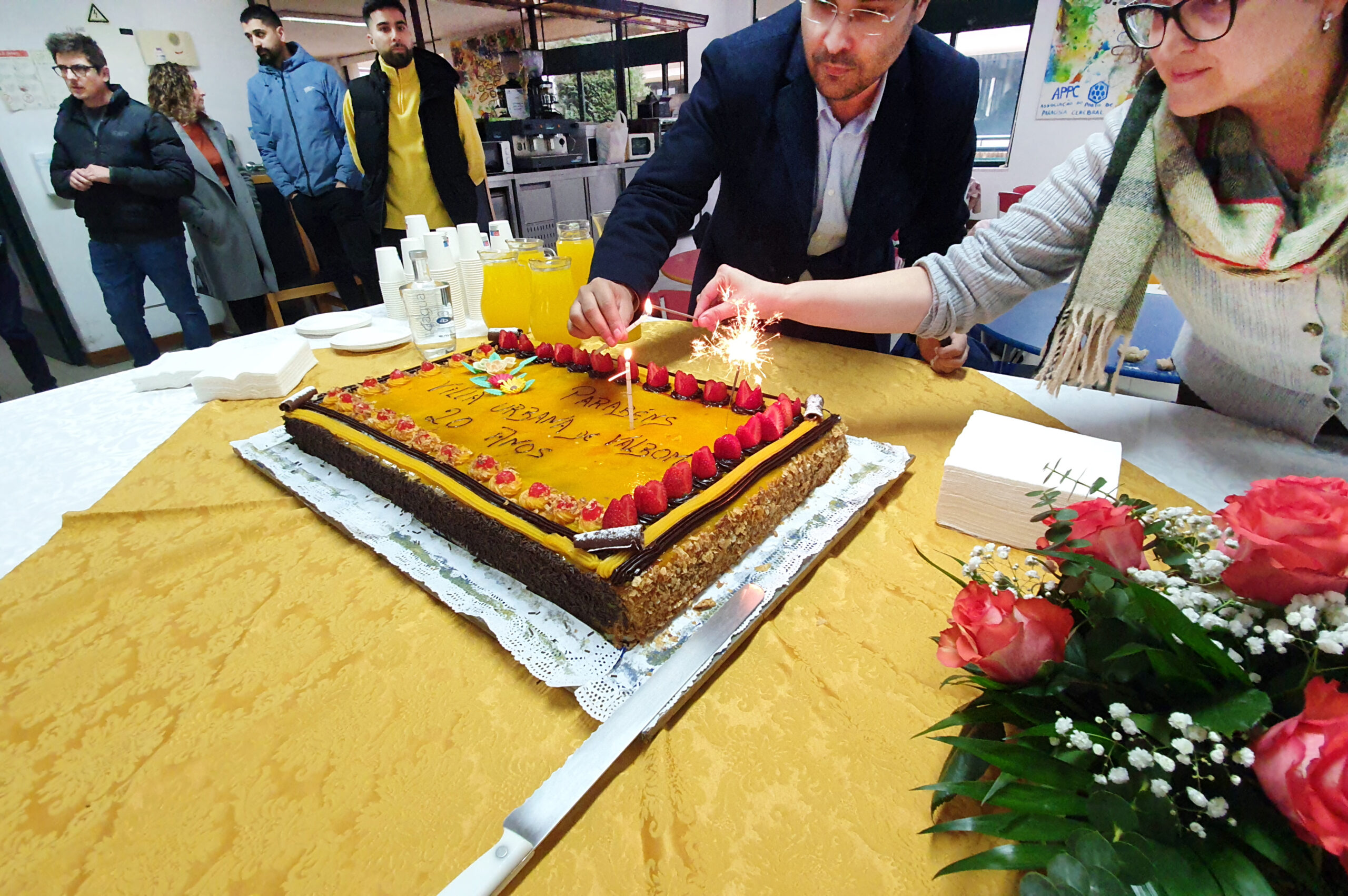 Duas pessoas acendem as velas de um bolo de aniversário. Em cima da mesa, além do bolo, está um arranjo floral, pratos, guardanapos, copos e jarros com sumo. Mais ao fundo da imagem outras quatro pessoas assistem.