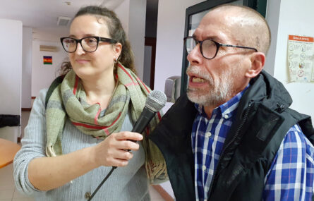 Fotografia com duas pessoas. Uma segura o microfone para a outra falar.
