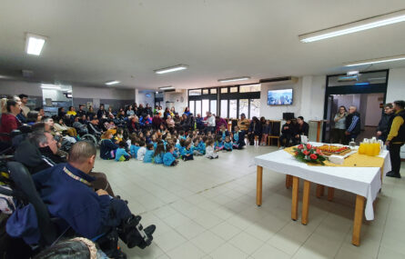 Várias dezenas de pessoas, adultos e crianças, em espaço interior da Villa Urbana de Valbom. À direita da imagem uma mesa com um bolo, um arranjo de flores, copos, pratos, guardanapos e vários jarros com sumo. Os rostos das crianças foram todos desfocados.