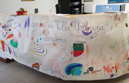 Fotografia da zona de entrada e de atendimento da Villa Urbana de Valbom. A secretária de atendimento está totalmente rodeada de um cartaz em tamanho gigante com várias mensagens e desenhos alusivos aos 20 anos da Villa Urbana – sendo que aparece, em destaque, a palavra Parabéns.