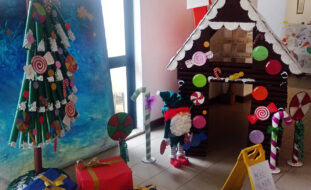 Fotografia de árvore de Natal com materiais reciclados, várias prendas no chão e, ao fundo, uma casa também com materiais reciclados.