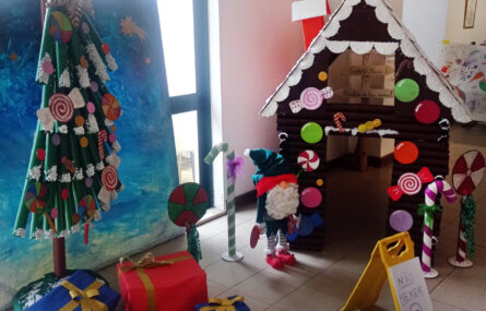 Fotografia de árvore de Natal com materiais reciclados, várias prendas no chão e, ao fundo, uma casa também com materiais reciclados.