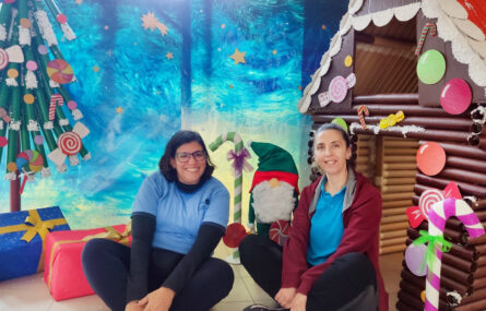 Duas pessoas sentadas, no chão, junto ao cenário elaborado (com árvore de Natal, prendas e casa de materiais reciclados).