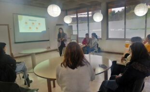 Fotografia de sala interior com sete pessoas sentadas. Ao centro uma mesa redonda e junto dessa mesa uma pessoa, em pé, a falar. Em painel na parede é feita a projeção de uma apresentação.