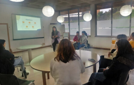 Fotografia de sala interior com sete pessoas sentadas. Ao centro uma mesa redonda e junto dessa mesa uma pessoa, em pé, a falar. Em painel na parede é feita a projeção de uma apresentação.