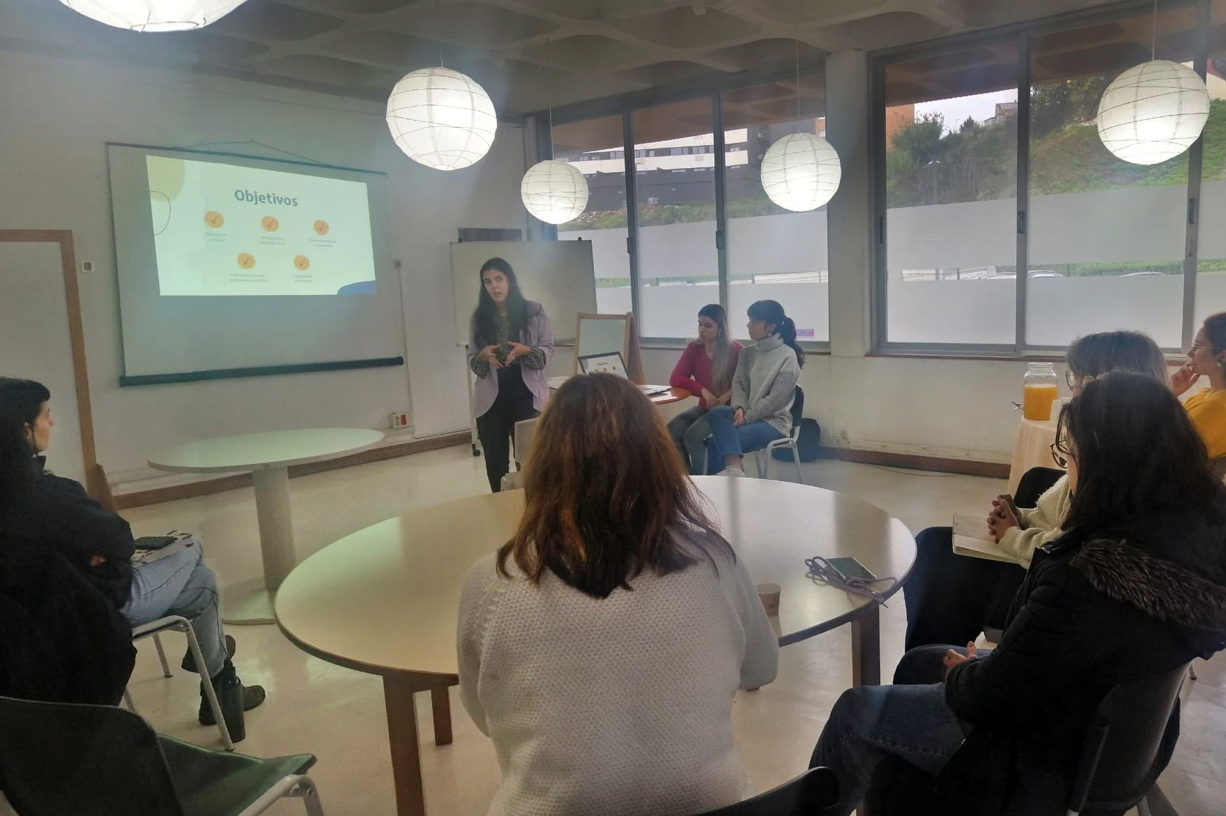 Fotografia de sala interior com sete pessoas sentadas. Ao centro uma mesa redonda e junto dessa mesa uma pessoa, em pé, a falar. Em painel na parede é feita a projeção de uma apresentação.