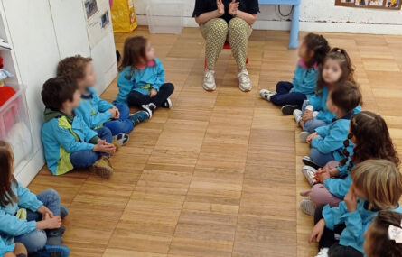 Grupo de 14 crianças, sentadas no chão de uma sala, a ouvir uma pessoa (sentada em pequena cadeira) a conversar com elas. Todos os rostos das crianças foram desfocados.