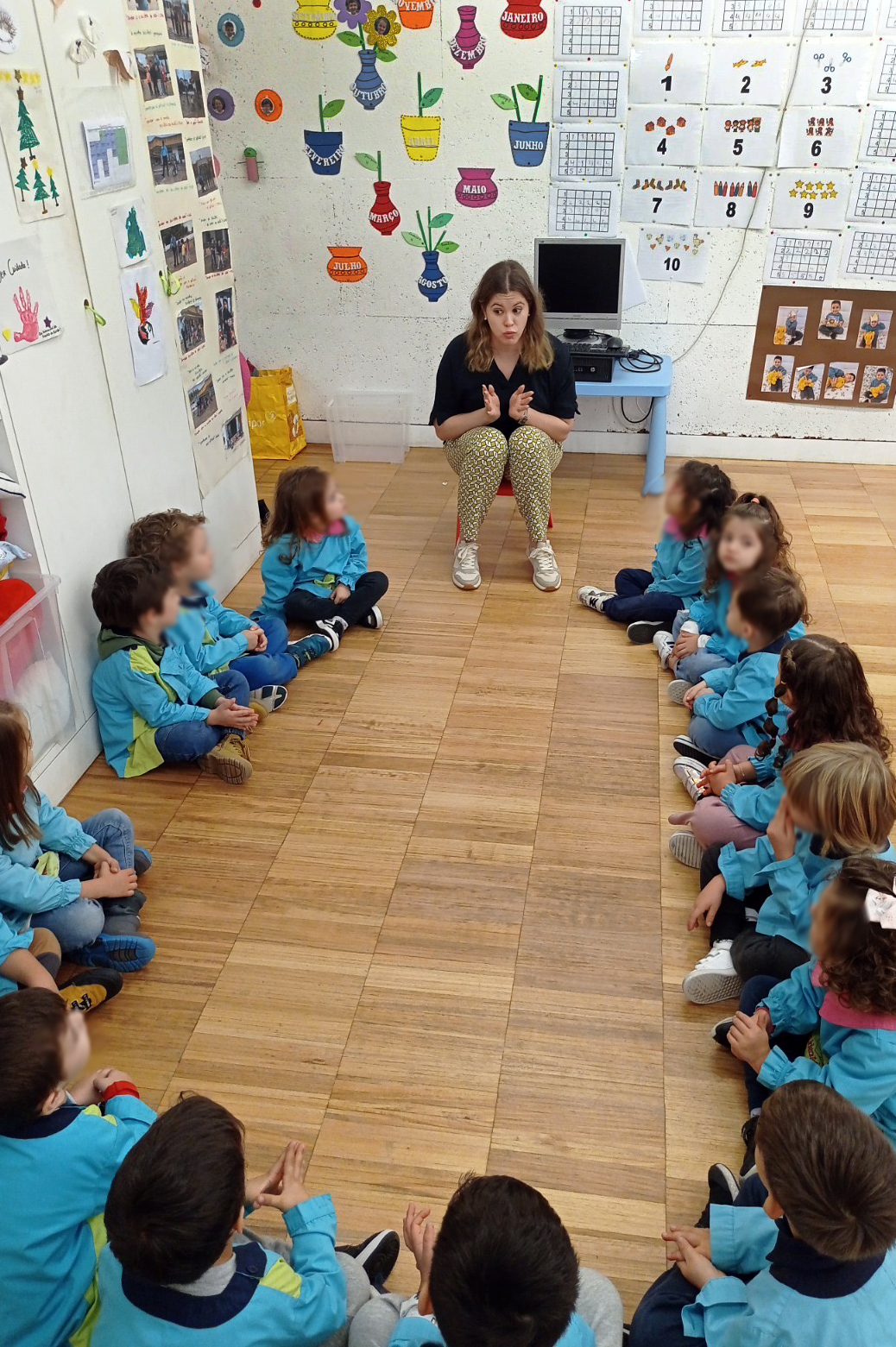 Grupo de 14 crianças, sentadas no chão de uma sala, a ouvir uma pessoa (sentada em pequena cadeira) a conversar com elas. Todos os rostos das crianças foram desfocados.