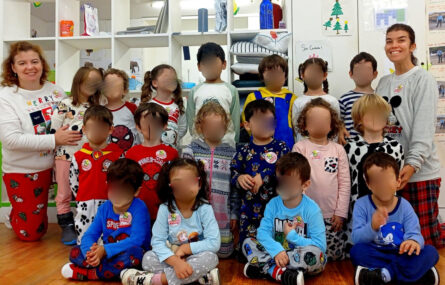 Grupo de 17 crianças, todas a usar pijama. Na fotografia aparecem ladeadas por duas pessoas adultas, também a usar pijama. Ao fundo da imagem prateleiras com diverso material. Todos os rostos das crianças foram desfocados.