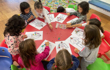 Grupo de nove crianças em redor de uma mesa com tampo vermelho. Na mesa estão várias folhas nas quais as crianças fazem a colagem de diverso material colorido. Todos os rostos das crianças foram desfocados.