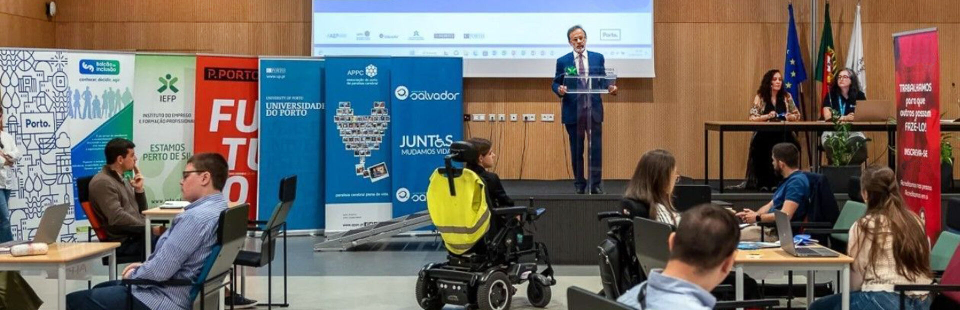 Fotografia de auditório. Ao fundo, em cima do palco, uma pessoa está a falar. Do lado esquerdo (da imagem) sete cartazes com identificação das entidades participantes na iniciativa – e já divulgadas no teor da publicação. Em primeiro plano, dos lados esquerdo e direito da imagem, várias pessoas sentadas junto de algumas mesas. Ao centro, junto do palco, uma pessoa sentada em cadeira de rodas.