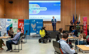 Fotografia de auditório. Ao fundo, em cima do palco, uma pessoa está a falar. Do lado esquerdo (da imagem) sete cartazes com identificação das entidades participantes na iniciativa – e já divulgadas no teor da publicação. Em primeiro plano, dos lados esquerdo e direito da imagem, várias pessoas sentadas junto de algumas mesas. Ao centro, junto do palco, uma pessoa sentada em cadeira de rodas.
