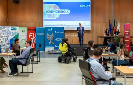 Fotografia de auditório. Ao fundo, em cima do palco, uma pessoa está a falar. Do lado esquerdo (da imagem) sete cartazes com identificação das entidades participantes na iniciativa – e já divulgadas no teor da publicação. Em primeiro plano, dos lados esquerdo e direito da imagem, várias pessoas sentadas junto de algumas mesas. Ao centro, junto do palco, uma pessoa sentada em cadeira de rodas.