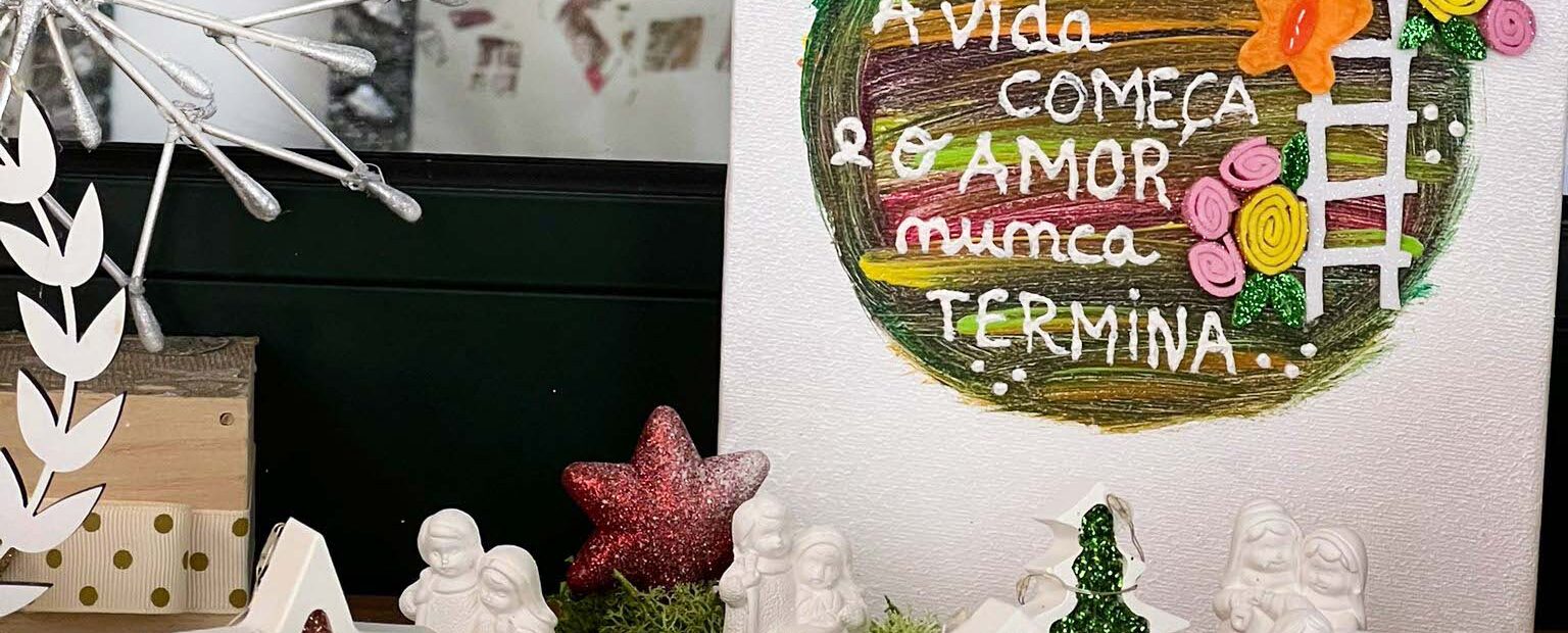Fotografia de arranjo de Natal em cima de uma mesa. Imagens de um presépio, duas estrelas e duas árvores. Atrás um cartaz com um círculo de várias cores e a mensagem: Família, onde a vida começa e o amor nunca termina.