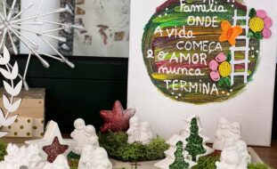 Fotografia de arranjo de Natal em cima de uma mesa. Imagens de um presépio, duas estrelas e duas árvores. Atrás um cartaz com um círculo de várias cores e a mensagem: Família, onde a vida começa e o amor nunca termina.