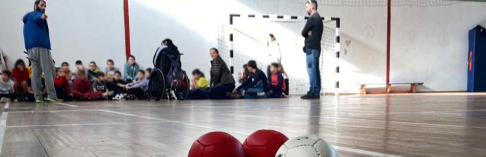 Imagem de interior de pavilhão desportivo. Em primeiro plano três bolas de Boccia. Ao fundo várias crianças e adultos. Os rostos das crianças foram todos desfocados.