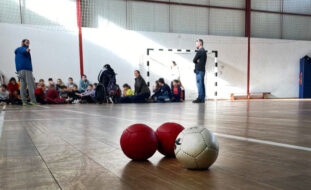 Imagem de interior de pavilhão desportivo. Em primeiro plano três bolas de Boccia. Ao fundo várias crianças e adultos. Os rostos das crianças foram todos desfocados.