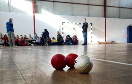 Imagem de interior de pavilhão desportivo. Em primeiro plano três bolas de Boccia. Ao fundo várias crianças e adultos. Os rostos das crianças foram todos desfocados.