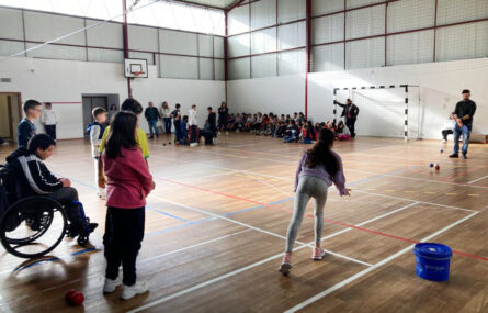 Imagem de interior de pavilhão desportivo. Em primeiro plano uma criança atira uma bola de boccia enquanto outras acompanham a jogada. Ao fundo várias crianças e adultos. Os rostos das crianças foram todos desfocados.