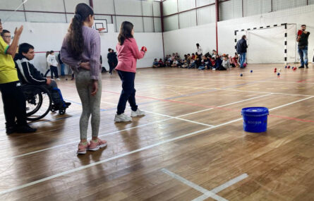 Imagem de interior de pavilhão desportivo. Em primeiro plano uma criança atira uma bola de Boccia enquanto outras acompanham a jogada. Ao fundo várias crianças e adultos. Os rostos das crianças foram todos desfocados.