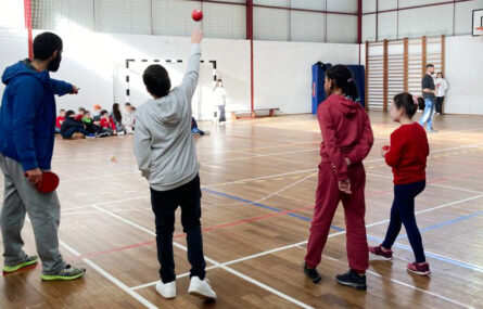 Imagem de interior de pavilhão desportivo. Em primeiro plano uma criança atira uma bola de Boccia enquanto outras acompanham a jogada. Ao fundo várias crianças e adultos. Os rostos das crianças foram todos desfocados.