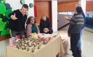 Mesa com vários produtos alusivos ao Natal. Atrás da mesa estão três pessoas. À frente da mesa, e a avaliar alguns produtos, estão dois possíveis compradores.