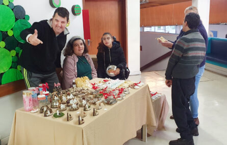 Mesa com vários produtos alusivos ao Natal. Atrás da mesa estão três pessoas. À frente da mesa, e a avaliar alguns produtos, estão dois possíveis compradores.