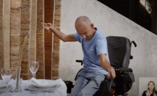 Captura de ecrã de vídeo. Pessoa em cadeira de rodas junto de uma mesa.