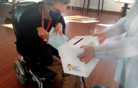 Pessoa em cadeira de rodas segura em envelope preparando-se para o colocar em urna de voto. A urna (com indicação “Voto antecipado Eleições Legislativas) está nas mãos de uma outra pessoa.